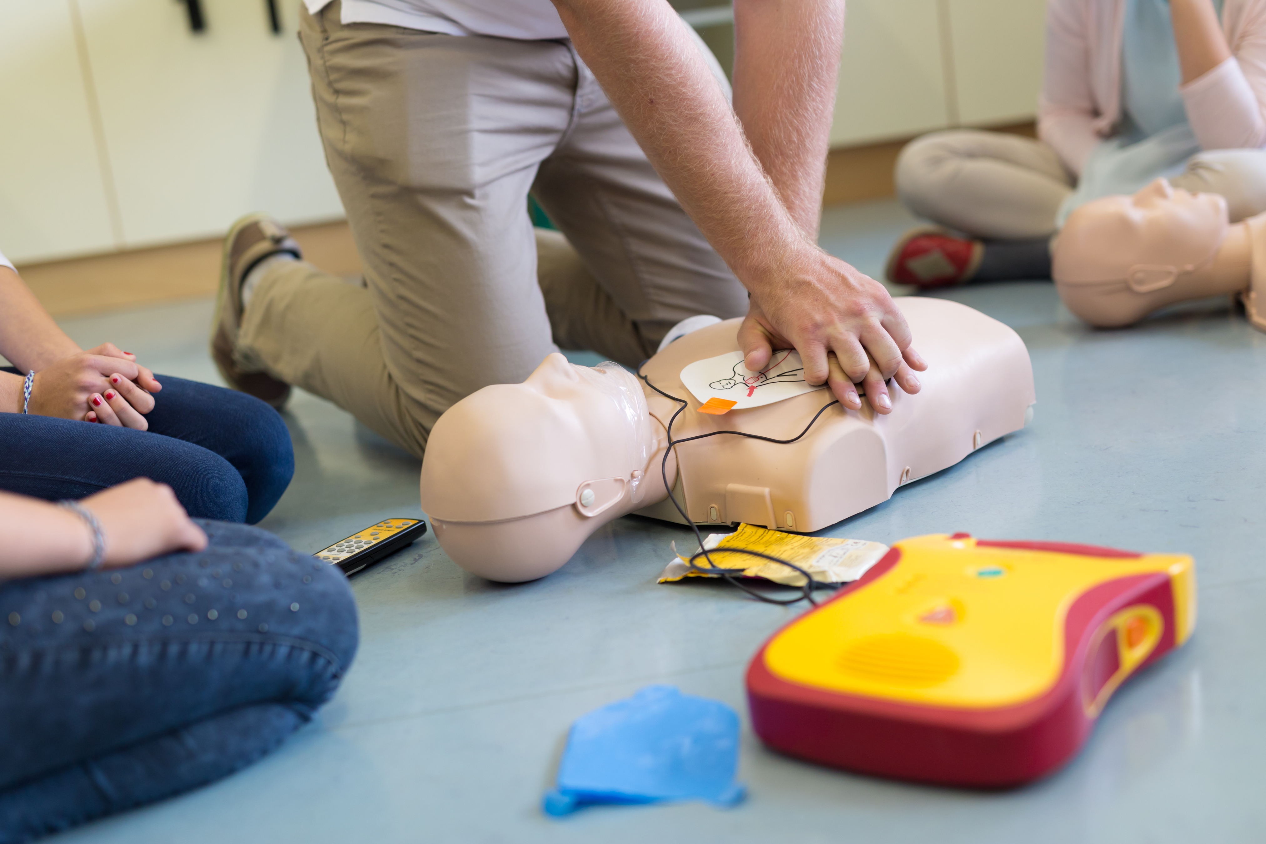 An individual practices CPR compressions on a CPR manikin, there is an AED pad in the foreground