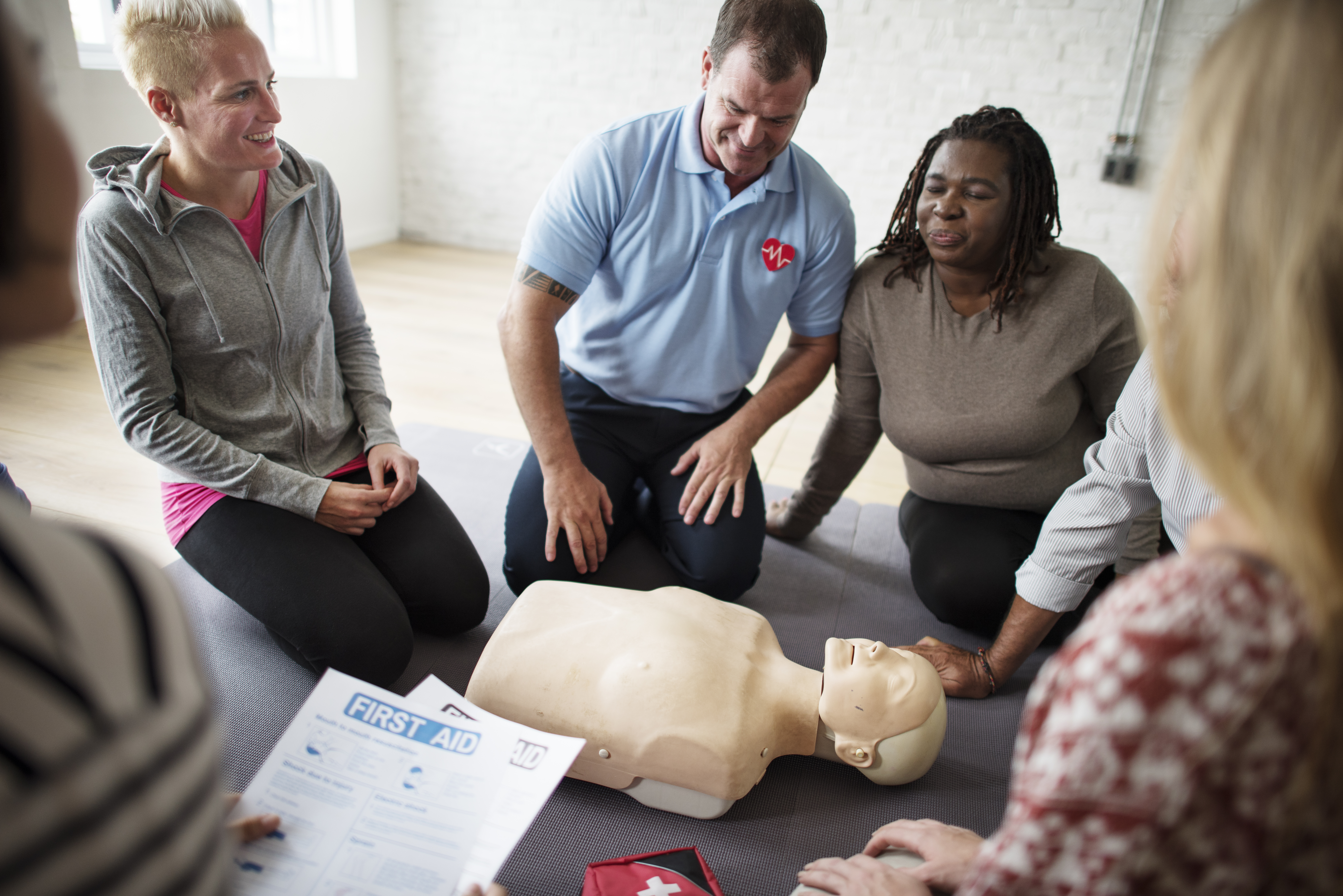 A diverse group of individuals gathers around a CPR mannequin with a Certified CPR Instructor. They are all smiling and engaged in the training.